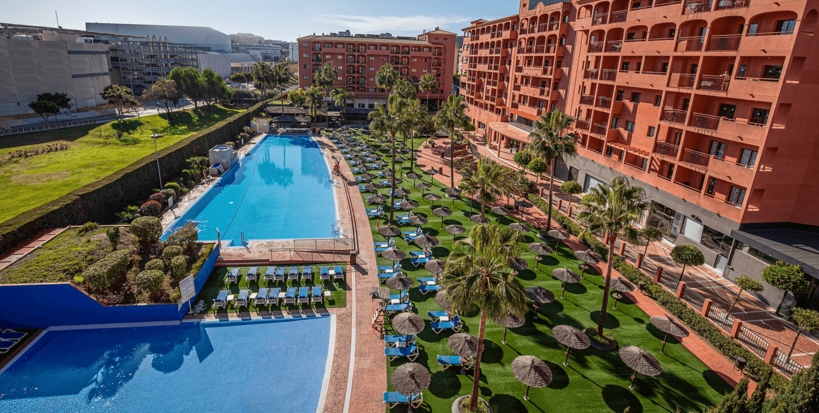 Aerial view of a terracotta resort with blue swimming pools, palm trees, and straw umbrellas.