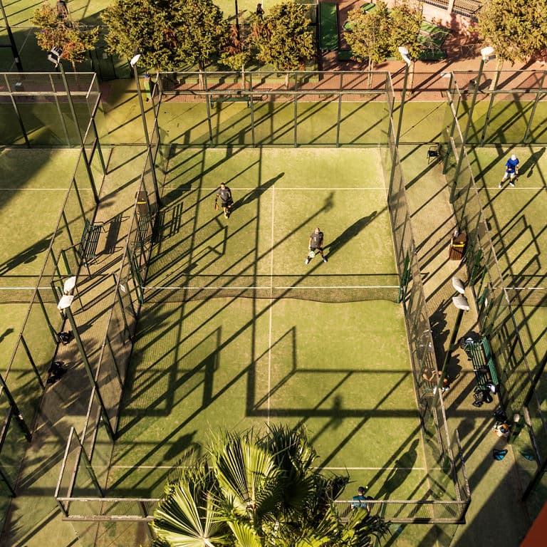 High-angle view of padel players on green courts with long shadows from the sun.